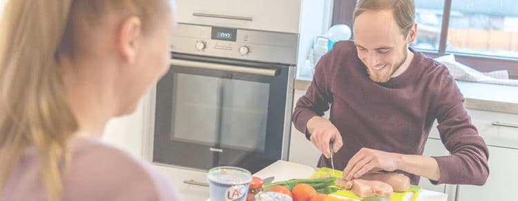 Junger Mann schneidet Fleisch und Gemüse, Freundin schaut dabei zu