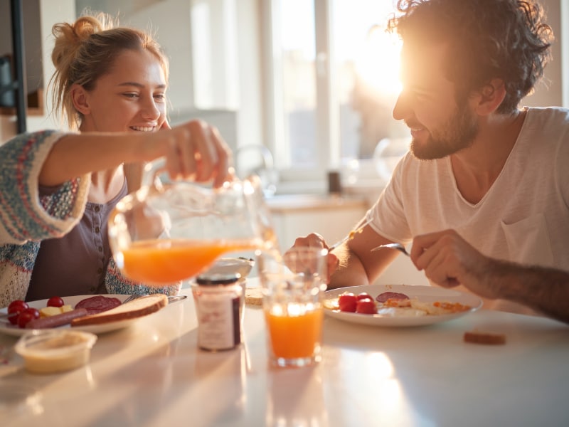 Glückliches Pärchen sitzt am Frühstückstisch, sie schenkt ihm Orangensaft ein