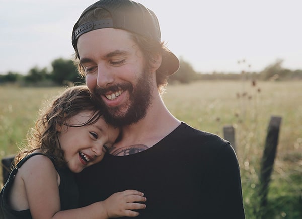 Junger Vater mit Bart und Baseball-Cap hält seine kleine lachende Tochter auf dem Arm