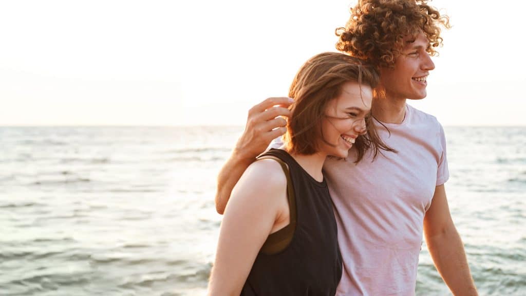 Pärchen spaziert in kurzen Sommerklamotten an einem Strand, im Hintergrund ist das Meer