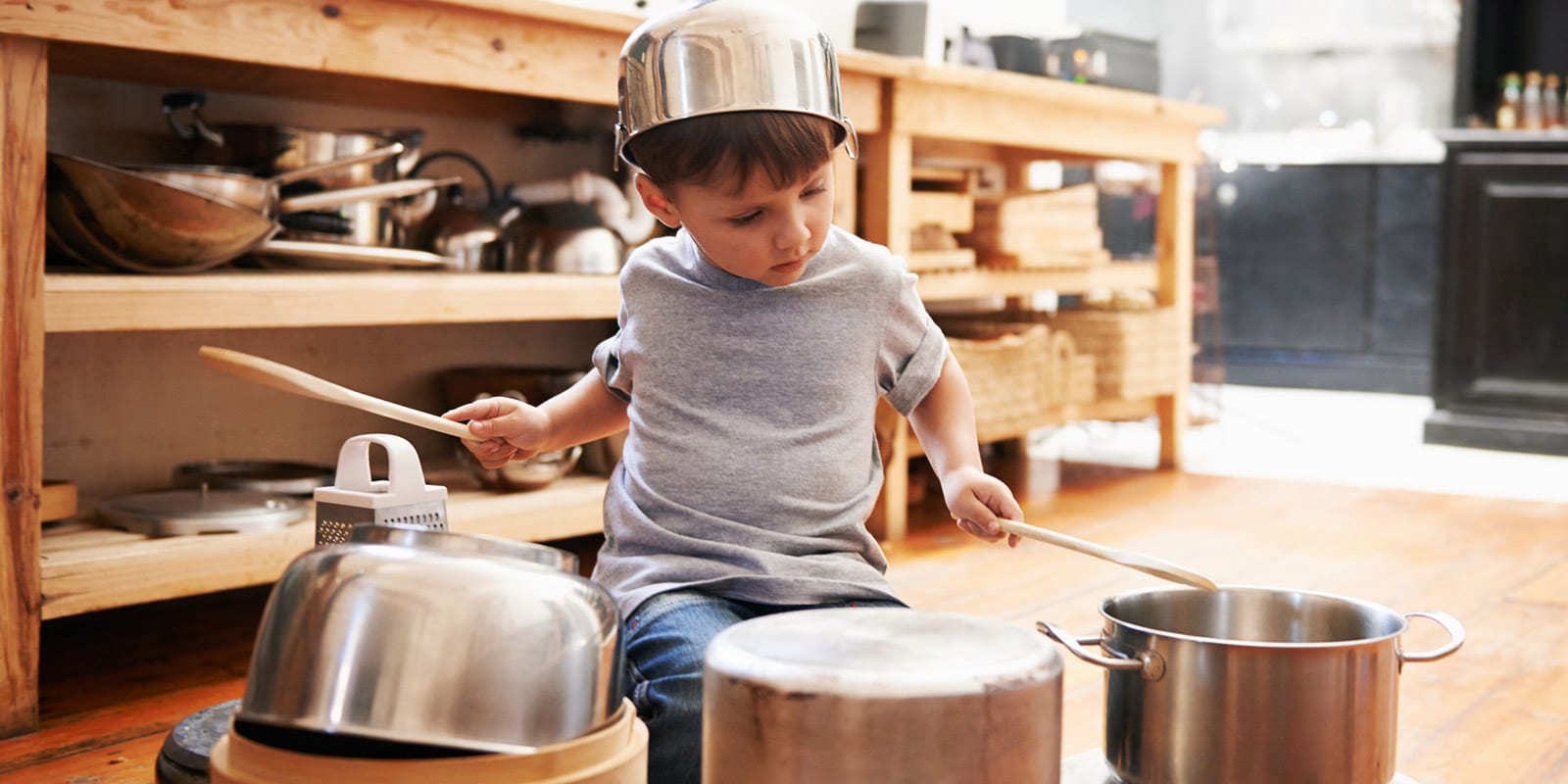 Ein kleiner Junge sitzt auf dem Küchenboden hält zwei Kochlöffel in den Händen und spielt auf Töpfen Trommel