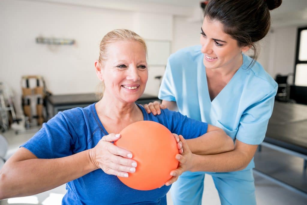 Eine junge Physiotherapeutin arbeitet mit einer älteren Patientin zusammen, die mit ihren Händen einen Ball zusammenpresst
