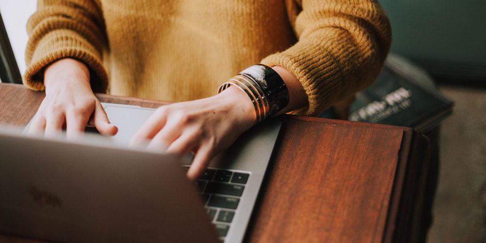 Frau mit senffarbenem Pullover sitzt am Schreibtisch und tippt auf der Tastatur ihres Laptops