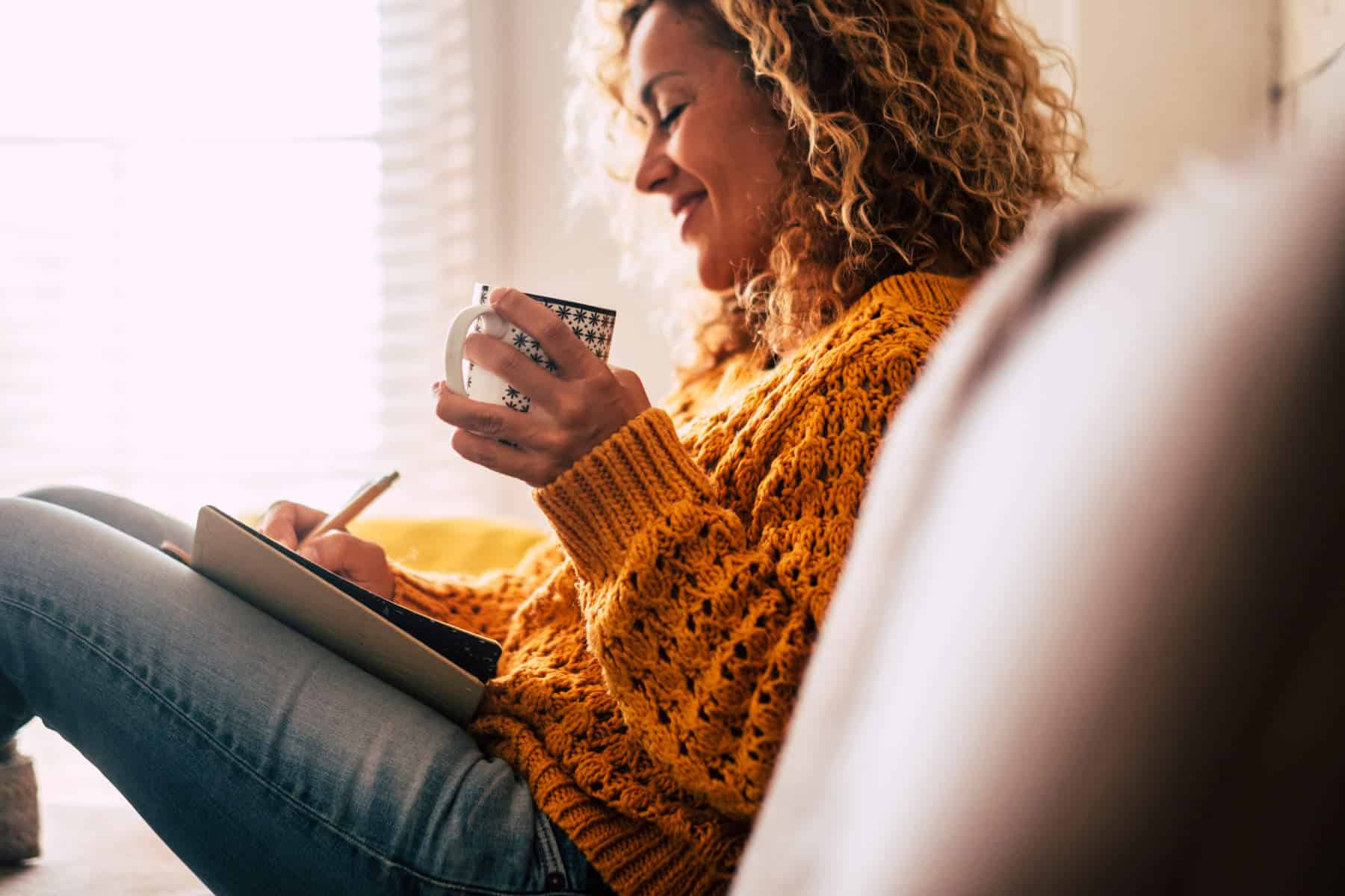 Frau mit lockigen Haaren sitzt auf dem Sofa und notiert etwas in einem Buch